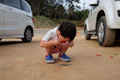 Boy photographing with toy camera while crouching by cars on field