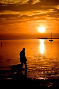 Silhouette man with dog standing in sea against sky during sunset