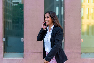 Smiling young woman using phone while standing against wall