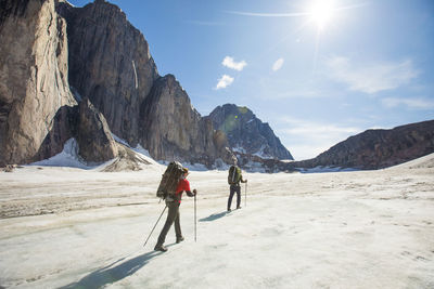 Two backpackers hiking on glacier below steep mountains.