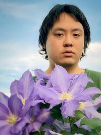 Close-up portrait of young man behind purple clematis flowers against blue sky.