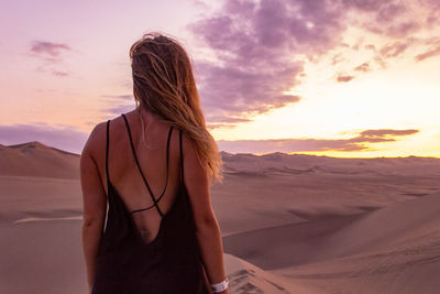 Rear view of young woman standing on sand dune against cloudy sky at sunset