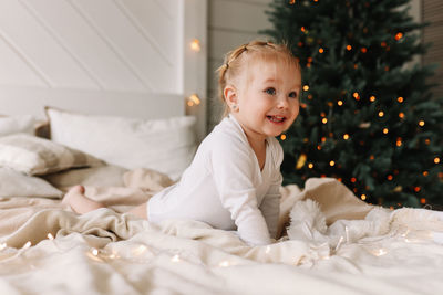Happy cheerful baby little girl sitting smiling on a cozy bed at christmas holidays at home
