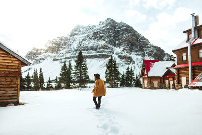 Back view of unrecognizable trendy female walking on show near lumber hut on shore of bow lake on winter day in alberta, canada