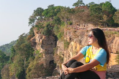 Woman wearing sunglasses while sitting on cliff against trees