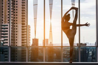 Rear view of young woman exercising against window in city during sunset