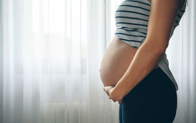 Midsection of pregnant woman touching her belly against curtain at home