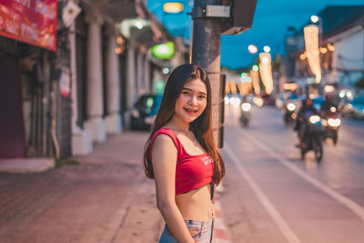 Portrait of smiling young woman standing on street in city at dusk