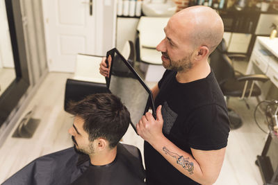 High angle view of bald barber holding mirror while showing to customer in salon