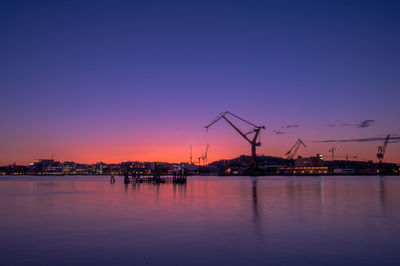 Silhouette cranes at commercial dock against sky during sunset