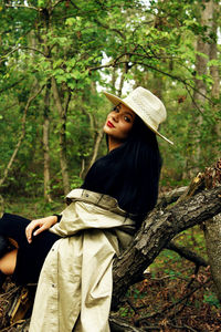 Portrait of young woman wearing hat while sitting on log in forest