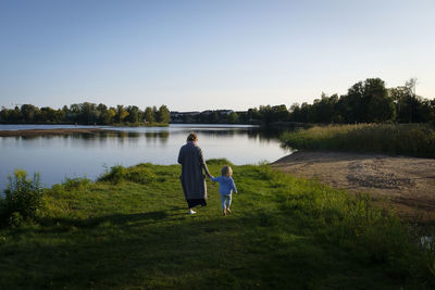 Rear view of mother and daughter walking by lake against sky