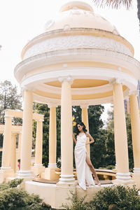 A beautiful brunette lady in an elegant wedding dress poses among the columns in the old city park