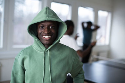 Portrait of smiling teenage boy in green hooded shirt standing in games room