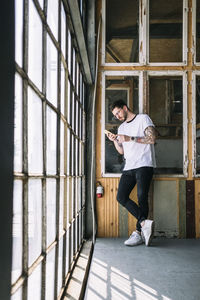 Full length of confident male hipster hacker using smart phone by window at creative office