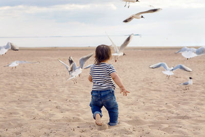 Baby boy in striped sailor t-shirt running on the sandy beach with seagulls near the sea in summer