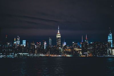 Illuminated buildings by river against sky at night