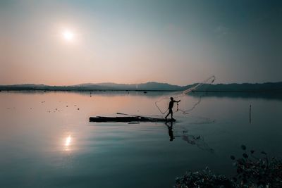 Scenic view of lake against sky with silhouette of a man casting net. 