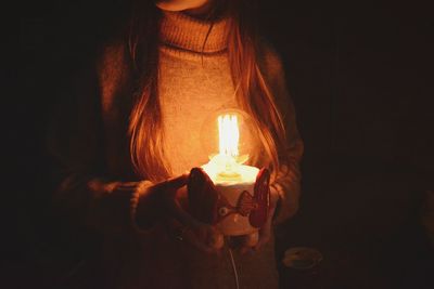 Midsection of woman holding illuminated light bulb while standing in darkroom