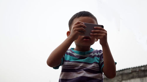 Low angle view of boy using phone while standing against clear sky