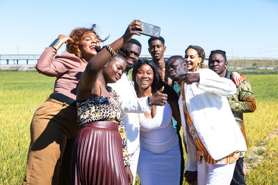 Happy african friends in stylish clothes smiling and taking selfie via cellphone in grassy field against blue sky together