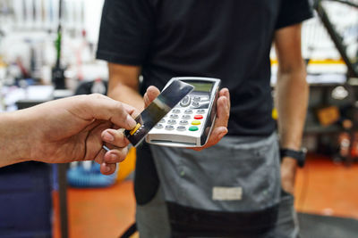 Low angle of crop client using smartphone to pay to male technician in repair workshop