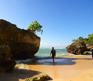 Rear view of men walking at beach by rock against clear blue sky