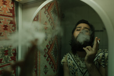 Bearded man smoking cigarette reflecting on mirror at home