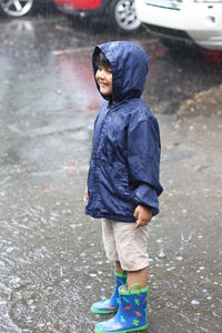 Full length of boy in raincoat looking away while standing on road during rainy season