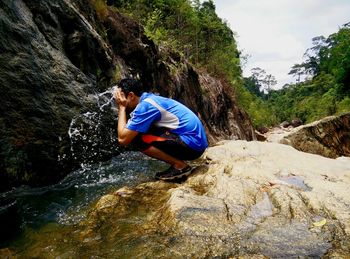 Man washing face with water by stream while crouching on rock