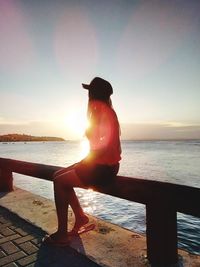 Man sitting on railing by sea against sky during sunset