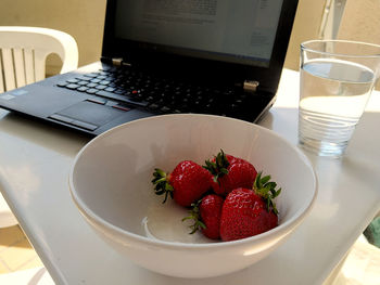 Strawberries in bowl on table with glass of water and a laptop