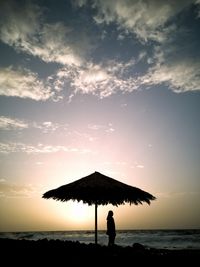 Silhouette woman standing by thatched roof parasol at beach during sunset