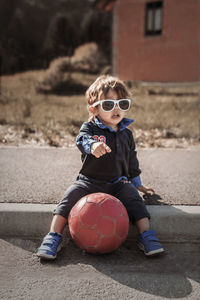 Full length of cute boy sitting with soccer ball on footpath