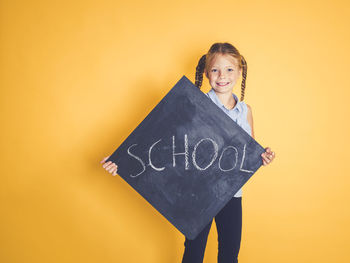 Portrait of smiling girl holding blackboard with school text while standing against yellow background