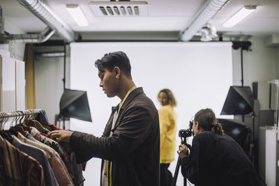 Side view of fashion designer choosing clothes from rack during photo shoot at studio