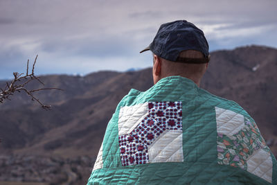 Rear view of man with blanket looking at mountains against sky