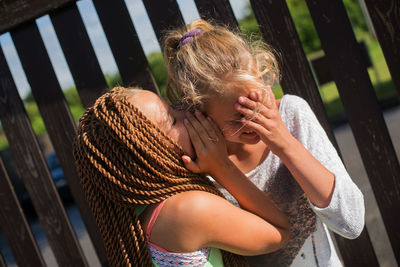 Tilt image of girl whispering to female friend by fence