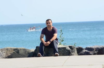 Portrait of man sitting on rock by promenade against sea