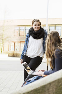 Happy schoolgirl looking at friend using laptop outside school
