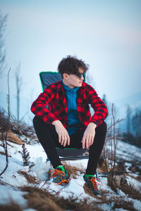 Handsome teenager aged 15-20 in a plaid red-black shirt and sunglasses on a chair by the tent.