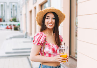Smiling beautiful asian young woman with long hair in straw hat drinking orange juice at city street