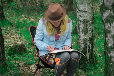 Young woman drawing in book by tree trunk
