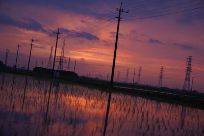 Silhouette electricity pylon by lake against romantic sky at sunset