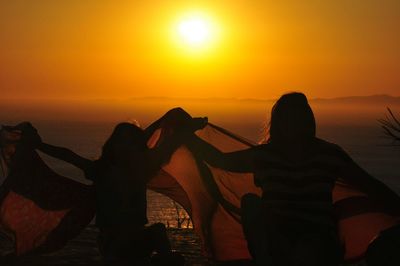 Mother and daughter holding fabric against sea during sunset