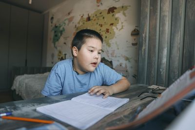 Asian boy reads during homework session with geography wall map, home education, e-learning