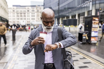 Senior businessman with disposable coffee cup checking time on wristwatch in city