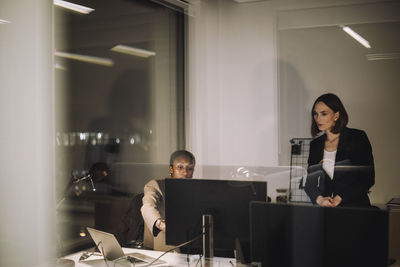 Female colleague discussing with businesswoman over computer seen through glass of office