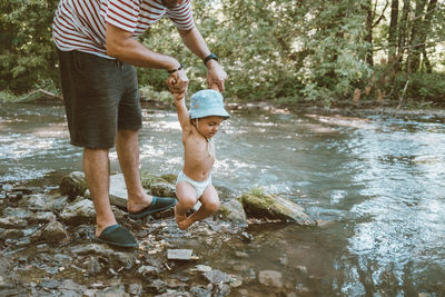 Low section of father holding baby boy hands over river in forest