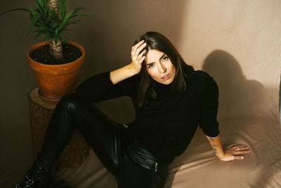 Young woman sitting by potted plant against wall at home